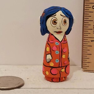 Puede incluir: Mu&ntilde;eca de madera pintada a mano de una ni&ntilde;a con cabello azul, vestida con un vestido de lunares rojos y blancos con detalles amarillos y azules. La mu&ntilde;eca mide aproximadamente 5 cm de alto.