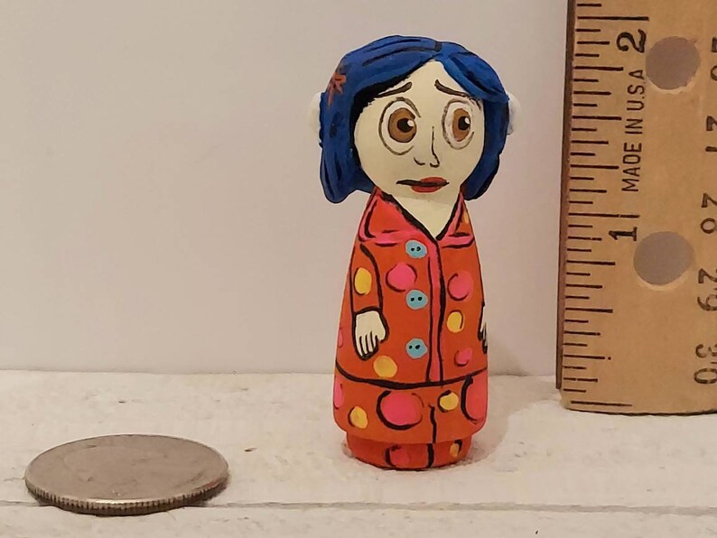 Puede incluir: Mu&ntilde;eca de madera pintada a mano de una ni&ntilde;a con cabello azul, vestida con un vestido de lunares rojos y blancos con detalles amarillos y azules. La mu&ntilde;eca mide aproximadamente 5 cm de alto.