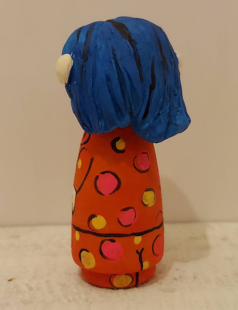 Puede incluir: Una mu&ntilde;eca de madera pintada a mano con cabello azul y un vestido rojo con lunares rosas y amarillos.