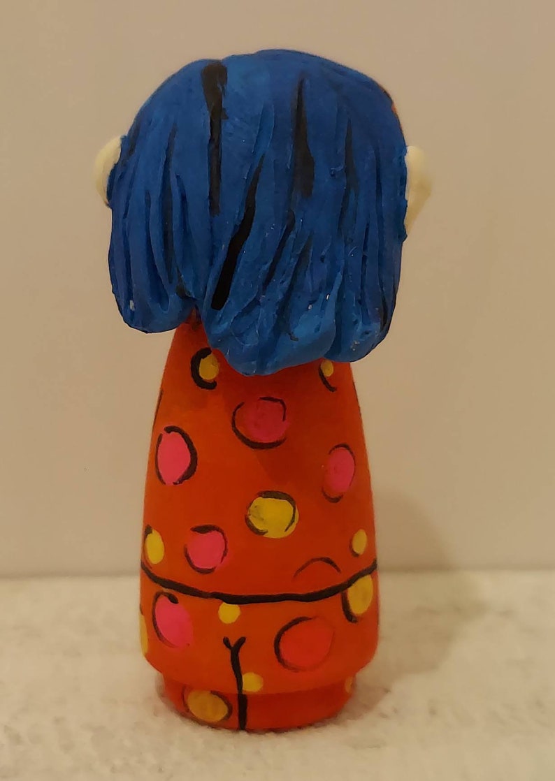 Puede incluir: Una mu&ntilde;eca pintada a mano con cabello azul y un vestido rojo con lunares rosas y amarillos.
