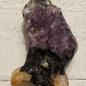 May include: A pendant necklace featuring a black resin hand with a cluster of purple amethyst crystals on top and three clear quartz crystals at the bottom. The pendant is attached to a brown leather cord.