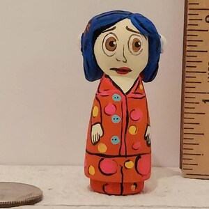 Puede incluir: Mu&ntilde;eca de madera pintada a mano con cabello azul, vestida con un vestido de lunares rojos y blancos. La mu&ntilde;eca tiene una cara blanca con ojos marrones y una expresi&oacute;n triste.