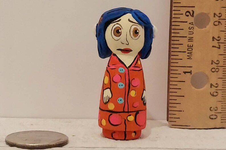 Puede incluir: Mu&ntilde;eca de madera pintada a mano con cabello azul, vestida con un vestido de lunares rojos y blancos. La mu&ntilde;eca tiene una cara blanca con ojos marrones y una expresi&oacute;n triste.