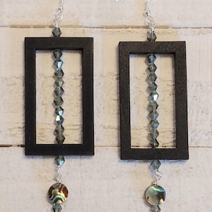 May include: A pair of handcrafted earrings featuring black rectangular frames with a vertical strand of faceted gray beads. Each earring has a dangling iridescent abalone shell bead and silver-tone ear wires. The earrings are displayed against a white wood-plank background.