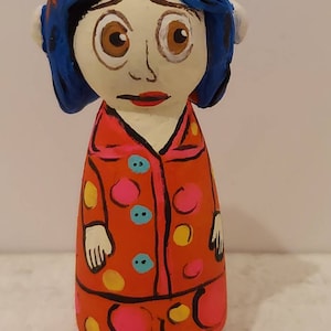 Puede incluir: Una mu&ntilde;eca pintada a mano de una ni&ntilde;a con cabello azul, vestida con un vestido de lunares rojos y rosados con botones amarillos y azules.