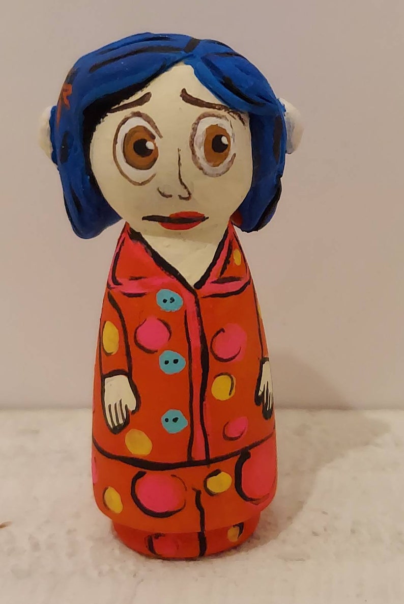 Puede incluir: Una mu&ntilde;eca pintada a mano de una ni&ntilde;a con cabello azul, vestida con un vestido de lunares rojos y rosados con botones amarillos y azules.