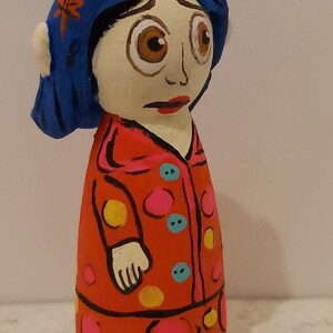 Puede incluir: Una figura de mu&ntilde;eca pintada a mano de una ni&ntilde;a con cabello azul, vestida con un vestido rojo con lunares amarillos y rosados. La mu&ntilde;eca tiene una expresi&oacute;n triste y mira hacia abajo.