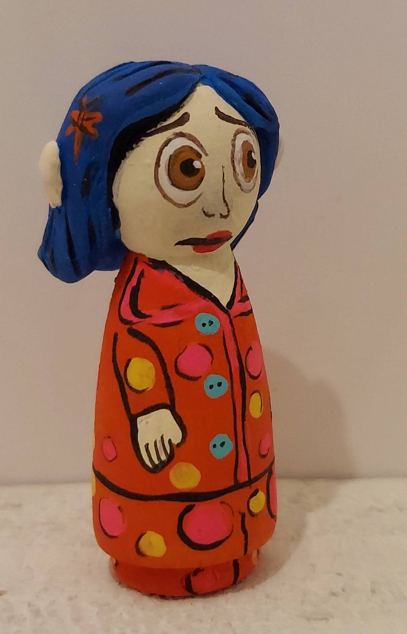 Puede incluir: Una figura de mu&ntilde;eca pintada a mano de una ni&ntilde;a con cabello azul, vestida con un vestido rojo con lunares amarillos y rosados. La mu&ntilde;eca tiene una expresi&oacute;n triste y mira hacia abajo.