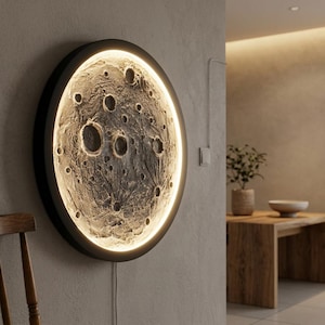 May include: A round wall-mounted light fixture resembling the moon. The fixture has a black frame and a textured surface with craters, lit by an internal light source. The light emits a warm glow, creating a celestial ambiance. A power cord extends from the fixture.