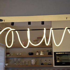May include: A modern, linear LED pendant light fixture with a black frame and a swirling, warm white neon light design. The fixture is suspended from the ceiling and illuminates a kitchen area with shelves and appliances.