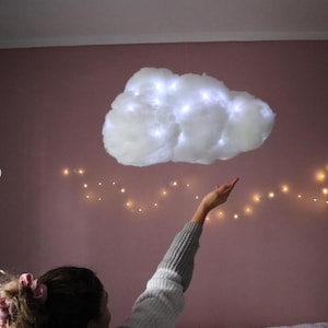 May include: A fluffy white cloud-shaped light fixture with embedded fairy lights, suspended against a pink wall. The cloud is positioned above a person's outstretched hand, with a string of warm white lights in the background. A whimsical decor item.
