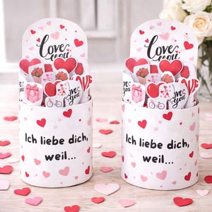 May include: Two white cylindrical containers decorated with red and pink hearts. Each container has a curved top with the word "Love" and "Love you" in script. The containers are filled with heart-shaped cards and have the German text "Ich liebe dich, weil..."