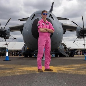 Puede incluir: Una persona con un mono rosa y gafas de sol está de pie frente a un gran avión militar gris. La persona tiene los brazos cruzados. El avión tiene cuatro hélices y está estacionado en una pista con líneas amarillas.