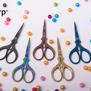 May include: Five pairs of CuT Sharp scissors in black, iridescent rainbow, bronze, gold, and blue finishes. Each pair features ornate, embossed handle designs and sharp blades. The scissors are arranged among scattered, colorful plastic beads.