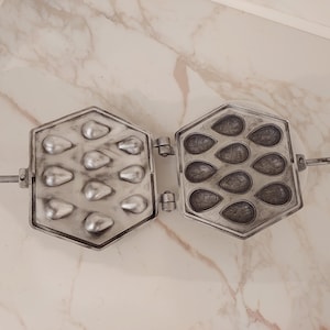 May include: Open, antique metal waffle iron with a hexagonal shape. The iron is silver-colored and has molds for making small, teardrop-shaped waffles. The iron is hinged and has a long handle for easy use. The background is a marble-patterned surface.