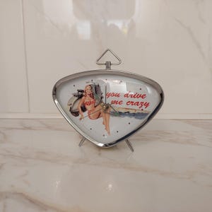 May include: A retro-style, silver-toned alarm clock with a triangular face. The clock face features a vintage pin-up girl illustration with the text "you drive me crazy". The clock has a small handle at the top and three legs.
