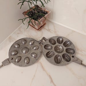 Könnte beinhalten: Ein antikes Waffeleisen aus Metall mit zwei runden Abschnitten. Jeder Abschnitt hat acht einzelne Formen in Form eines Eies. Das Waffeleisen ist geöffnet und zeigt das Innere der Formen.