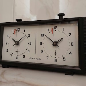 May include: A black chess clock with two white clock faces. Each clock face has black numbers and hands, with a red second hand. The clock has two black knobs on top and the word "Янтарь" is printed on the bottom.