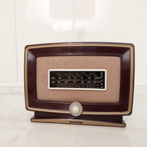 Puede incluir: Una radio Marconi vintage con un marco marrón oscuro y una cubierta de altavoz beige. La radio tiene un panel de control blanco con indicadores de frecuencia y un dial de sintonización. La base y el borde tienen un acabado dorado. La radio es de estilo retro.