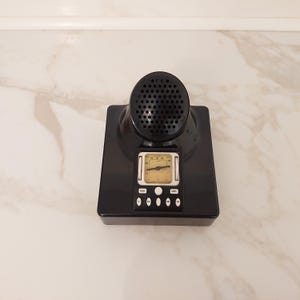 May include: A black plastic tabletop radio with a prominent circular speaker grille at the top. Below the speaker is a rectangular analog frequency display with a needle, flanked by two vertical silver bars. Five small white buttons are below the display.