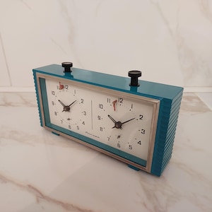 May include: A vintage teal chess clock with two white clock faces, black hands, and a silver frame. The clock has two black knobs on top and is designed for competitive chess play. The clock's face displays the numbers 1 through 12.