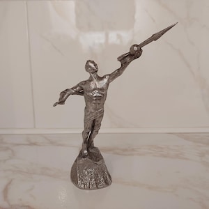 Può includere: Una statuetta metallica color argento che raffigura una figura in piedi dalla corporatura muscolosa, drappeggiata con un panno intorno alla vita. La figura tiene un oggetto simile a una lancia verso l'alto con un braccio alzato su una base rocciosa.
