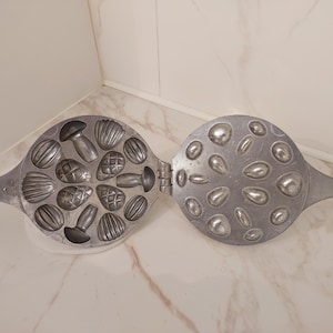 May include: Open, silver-colored, hinged mold for making shaped cookies or candies. One side features various shell, mushroom, and pinecone shapes. The other side has a variety of oval and teardrop shapes. The mold is open, revealing the interior.