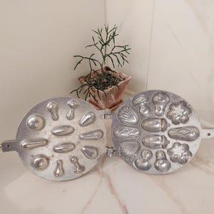 Puede incluir: Hierro para gofres vintage plateado abierto, con varios moldes con forma de alimentos, incluyendo setas, conchas y bellotas. El hierro está abierto, revelando el interior detallado. Una pequeña planta en maceta está en el fondo.