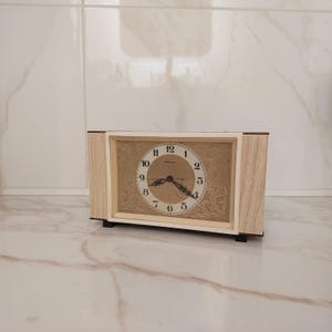 Pode incluir: Um relógio retangular vintage com moldura de madeira clara e mostrador creme. O mostrador do relógio apresenta algarismos e ponteiros pretos, com um design dourado ornamentado no fundo. O relógio tem aproximadamente 18 cm de largura.