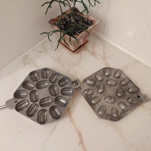 May include: A vintage metal waffle iron with two separate sections, each with 12 individual molds shaped like small, elongated ovals. The iron is made of a silver-colored metal and has a slightly worn appearance.