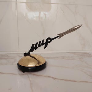 May include: A decorative sculpture featuring a gold globe base with a black rim. A black script reads "мир" with a silver rocket extending from it. The sculpture is set against a neutral background.