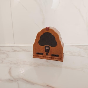 May include: A small, arched, light brown radio with a faux wood grain finish. It features a dark brown, cloud-shaped speaker grille, a central rectangular dial with white markings, and two horizontal black control knobs at the base.