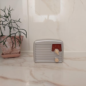 Könnte beinhalten: Ein silberfarbenes Radio im Retro-Stil mit rotem Zifferblatt und cremefarbenen Knöpfen. Das Radio steht auf einer Marmoroberfläche neben einer Topfpflanze. Das Radio hat ein klassisches, Vintage-Design.