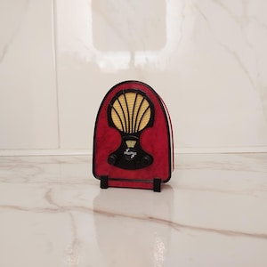 May include: A small, red and black vintage radio with a gold speaker cover. The radio features two black knobs and a dial. The design is reminiscent of mid-20th-century radios. The radio is approximately 15 cm tall.
