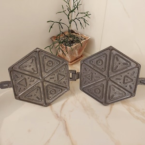May include: A vintage, cast-iron waffle iron with a hexagonal shape, featuring a geometric design of triangles and circles. The waffle iron is open, revealing the cooking surfaces. A small potted plant sits in the background.