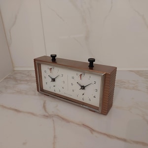 Puede incluir: Un reloj de ajedrez vintage con un marco de madera marrón y dos esferas blancas. Cada esfera muestra números y manecillas negras, con perillas negras en la parte superior. El reloj está diseñado para el juego de ajedrez competitivo.