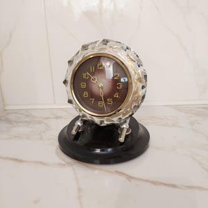 Puede incluir: Un reloj redondo vintage con una carcasa de cristal facetado y una esfera marrón oscuro. El reloj tiene manecillas doradas y una base negra. La esfera del reloj muestra los números del 1 al 12.