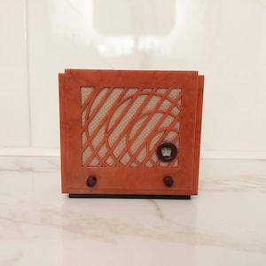 Op de afbeelding: Een radio in vintage-stijl met een warme oranjebruine afwerking. De voorkant heeft een geweven luidsprekercover met een geometrisch patroon. Het heeft twee donkere knoppen en een ronde wijzerplaat. De radio staat op een zwarte basis.