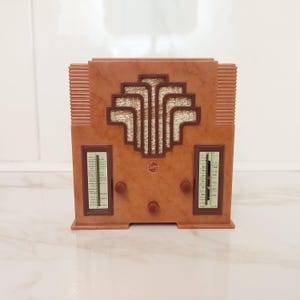 May include: A vintage-style radio in shades of brown and tan. The radio features an Art Deco design with a central geometric pattern and two vertical dials. It has a small, circular emblem and several knobs. The radio is a decorative item.
