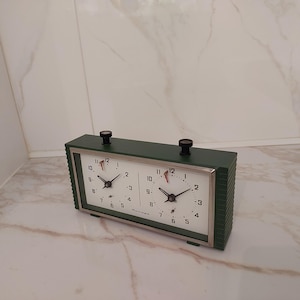 May include: A vintage green chess clock with two white clock faces. Each clock face has black numbers and hands, with a small black knob on top. The clock is set on a marble surface.