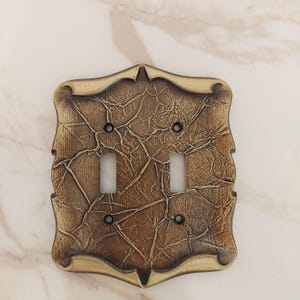 May include: A rectangular double light switch wall plate featuring a metallic bronze finish with a textured, cracked, or crinkled surface pattern. The plate has a decorative, scalloped edge design and includes four small screw holes for mounting.