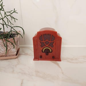 Op de afbeelding: Een kleine, roodbruine plastic radio met een gebogen bovenkant, een centrale luidsprekergrille met een decoratief uitgesneden patroon, een ronde afstemknop met een centrale draaiknop en twee kleinere bedieningsknoppen aan de voorzijde.