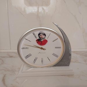 Può includere: Un orologio vintage con quadrante bianco, numeri romani neri e lancette dorate. L'orologio presenta l'immagine di un cosmonauta in uniforme rossa. L'orologio è posto su una base bianca e ha un accento argentato.