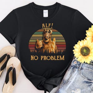 May include: Black t-shirt with a retro-style graphic of ALF, the alien from the 1980s television show. The graphic features ALF with the text "ALF! NO PROBLEM".