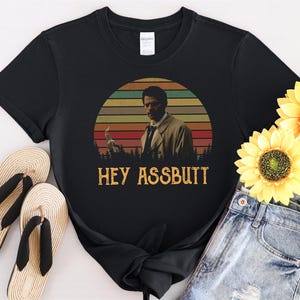 Könnte beinhalten: Schwarzes T-Shirt mit einem Retro-Sonnenuntergang-Motiv, auf dem ein Mann einen Feuerzeug hält. Der Text "Hey Assbutt" ist unter dem Motiv gedruckt.