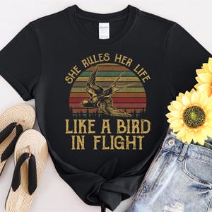 May include: Black t-shirt with a vintage-style graphic of a bird in flight against a sunset backdrop. The text reads "She Rules Her Life Like a Bird in Flight." The shirt is paired with denim shorts and sandals.
