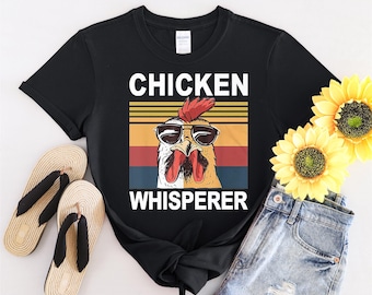 May include: Black t-shirt with a retro-style graphic of a chicken wearing sunglasses and the text "Chicken Whisperer".