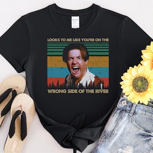 Puede incluir: Camiseta negra con un gráfico vintage de un hombre con el texto "Looks to me like you're on the wrong side of the river."