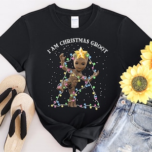 May include: Black t-shirt with a Groot graphic, decorated with Christmas lights and a star, and the text "I am Christmas Groot". The shirt is styled with denim shorts and sandals. The image also includes sunflowers.