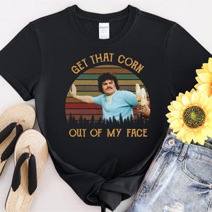 Może przedstawiać: Czarny T-shirt z retro grafiką mężczyzny z tekstem "Get That Corn Out Of My Face".